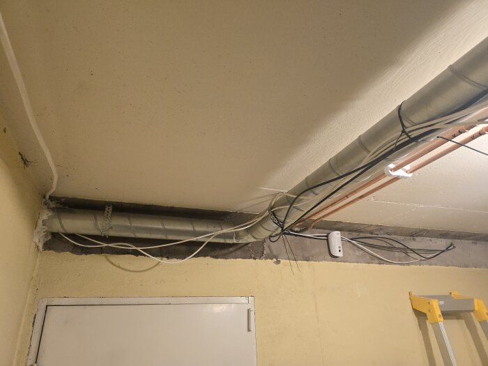 Tak med ventilationsrör och kablar, del av en renovering. En dörr och en stege syns mot en gul vägg i ett förråd.