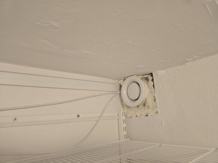 Ventilationsrör i ett hörn av vitmålad vägg med kablar och en hylla.
