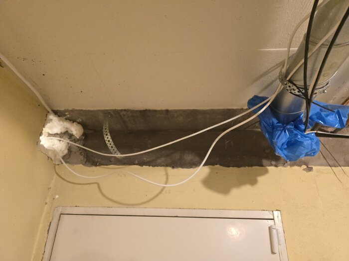 Ventilationsrör med kablar och blå plast, monterat i taket med isolering runt, ovanför en dörr i ett förråd.