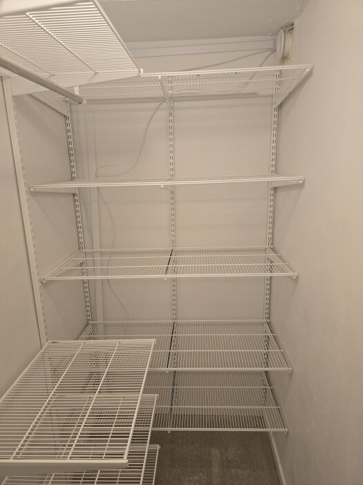 Tomt förråd med vita metallhyllor från Elfa, en synlig kabel och ett ventilationsrör i hörnet.