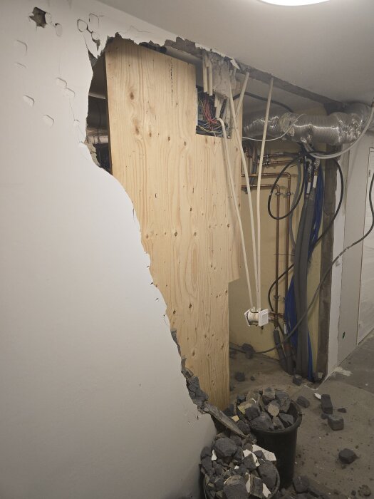 Riven innervägg med kabeldragning och ventilationsrör synliga. Brädhög på golvet i ett förråd under renovering.