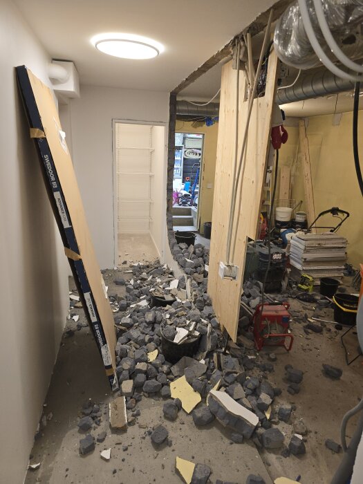 Renoveringsprojekt med nerbryten vägg, stenar och rivningsmaterial på golvet, verktyg och stege i bakgrunden, öppet förråd.