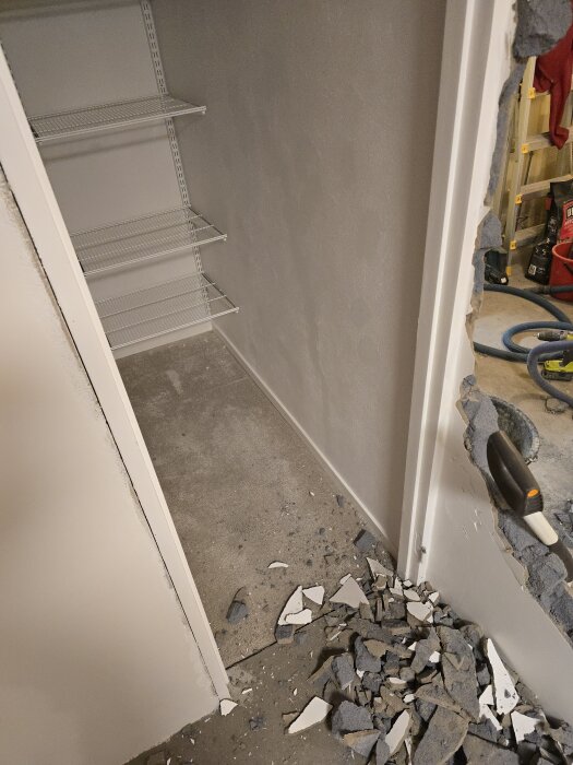 Förråd med trasig vägg, nedrivna bitar på golvet, tre metallhyllor på vänster sida, och verktyg i bakgrunden.