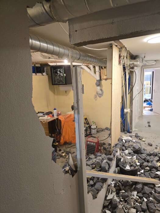 Renovering av förråd, halvdemonterad vägg med rör och ventilationssystem synliga. Skräp och bråte på golvet, arbete pågår.