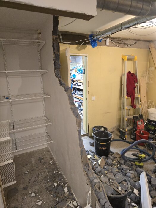 Riven vägg i rum, stenar på golvet, elfa-hyllor till vänster, stege och verktyg i hörnet. Ventilationsrör och kablar i taket.