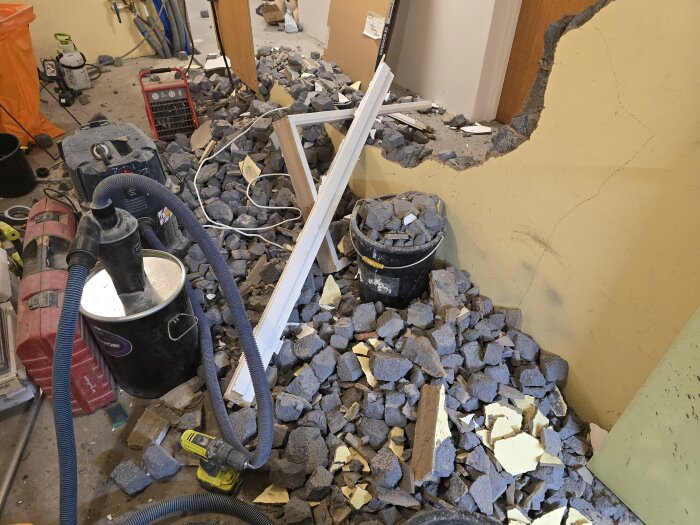 Rivningsprojekt med stenar och skräp från en nedriven vägg. Verktyg och sladdar synliga, med hinkar och dammsugare. Öppning i väggen under renovering.