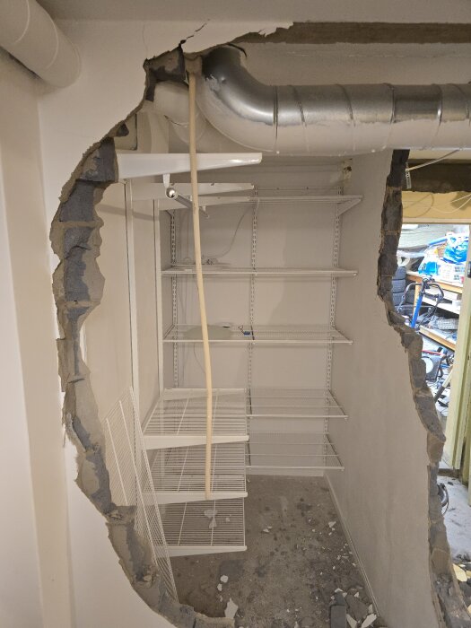 Riven vägg med synliga Elfa-hyllor, ventilationsrör och spillror på golvet. Förrådet omvandlas för ny planlösning.