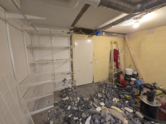 Rum under renovering med krossade betongrester på golvet, vita hyllor på väggarna, en stängd dörr och ventilationsrör i taket.