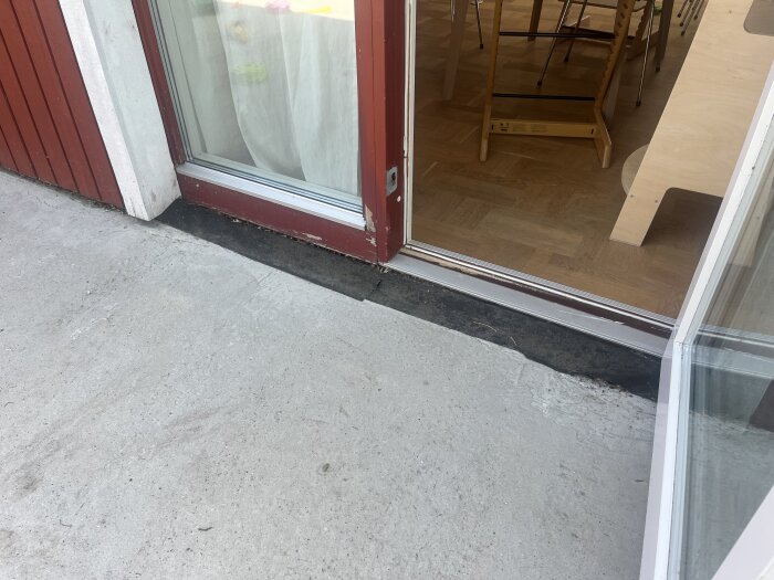 Utomhusbild på en altandörr med sliten tröskel och oregelbunden betongyta, inomhus syns parkettgolv och möbler.