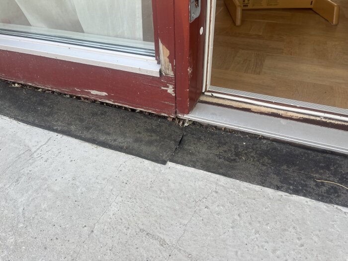 Dörrkarm i rött trä med färgavskavningar, synlig betongtröskel och svart matta synlig i förgrunden.