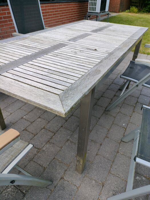 Slitet altanbord i trä med två stolar på stenlagd uteplats, omgivningen visar gräs och tegelvägg. Träet har flisor och behöver ytbehandling.