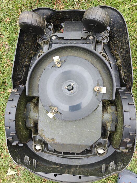 Upp-och-nervänd robotgräsklippare med gräsklippingsrester på undersidan, hjul och klipplatta synliga.