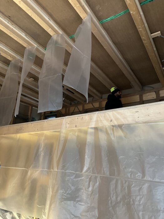 Byggprojekt under arbete, oskyddade takbjälkar och plastfolie hänger ner från taket, person i byggmiljö.