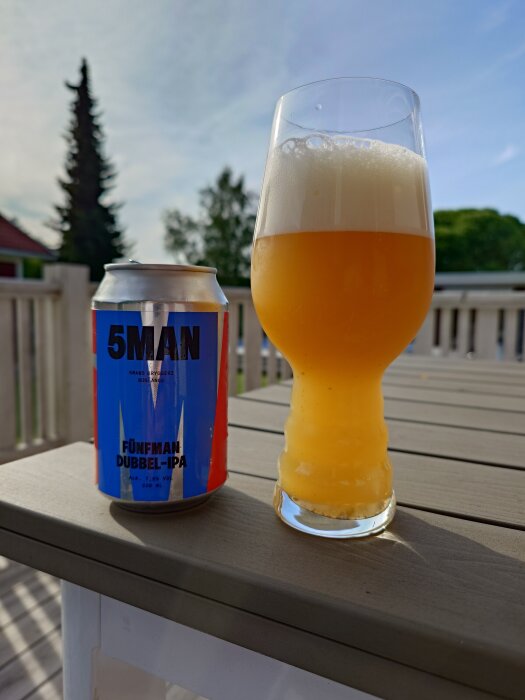 Burk med 5Mans Fünfman Dubbel-IPA bredvid ett glas öl på ett bord utomhus, med suddig bakgrund av träd och blå himmel.