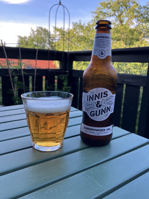 Flaska och glas med Innis & Gunn Gunnpowder IPA på ett grönmålat bord utomhus med träd och himmel i bakgrunden.
