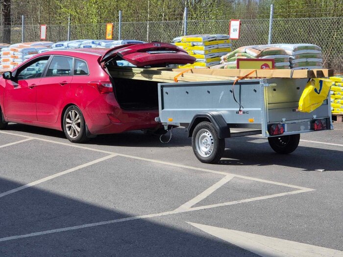 Röd bil med öppet bagage och släp fullt med byggmaterial, släpet verkar inte orka med lasten.