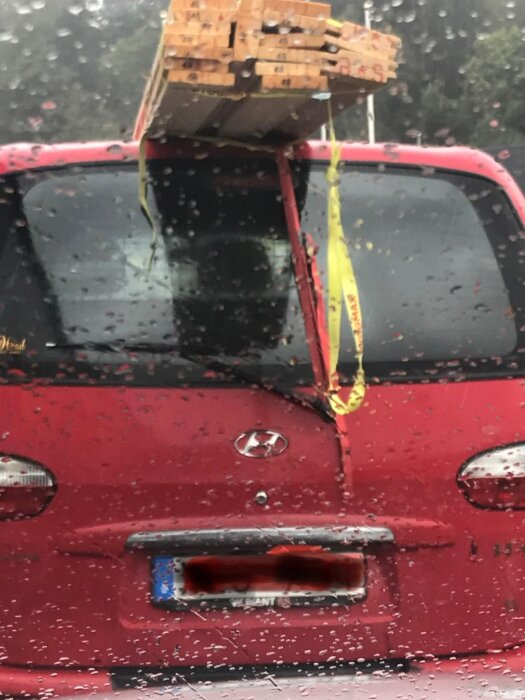 En röd bil med träplankor lastade på taket, fastsurrade med gula spännband. Plankorna sticker ut bakom bilen. Regn på bilrutan.