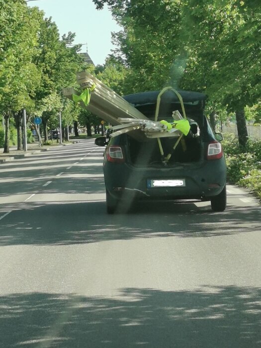 Svart bil med öppen baklucka, lastad med långa träplankor som sticker ut och säkrade med spännband, kör på en trädbevuxen väg.