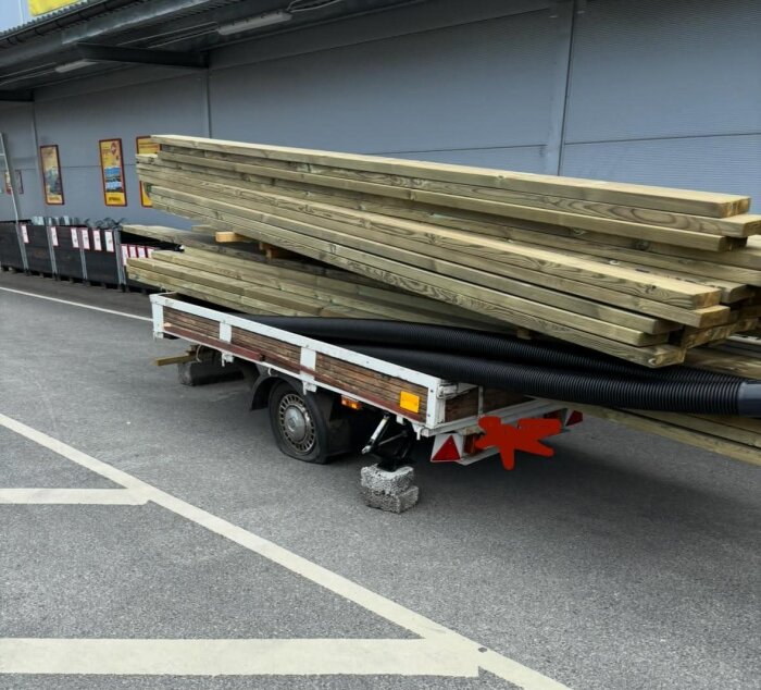 Släpvagn med nedtyngd axel, lastad med träbalkar och svarta rör, stöttad av betongblock på asfalterad yta.