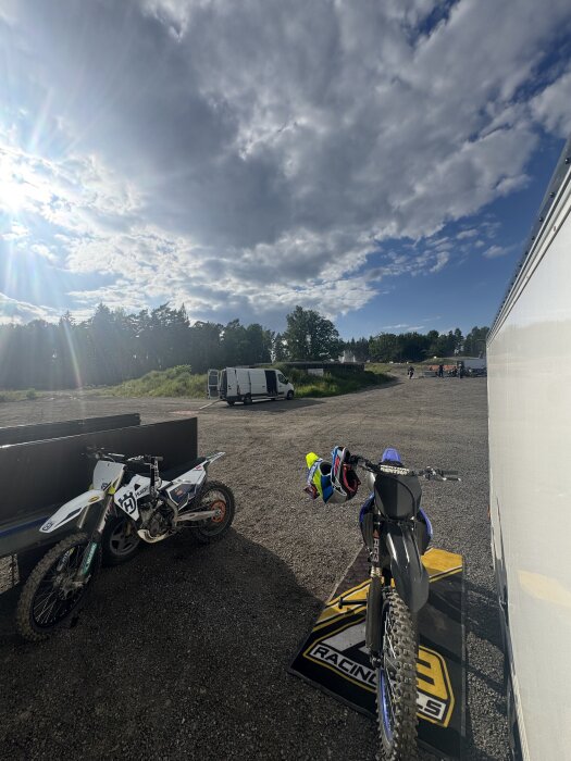 Motorcyklar parkerade på grusplan vid solig dag med molnig himmel; en vit Husqvarna och en svart motorcykel synliga bredvid en vit skåpbil.