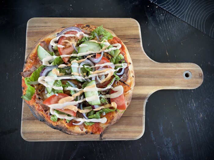 Kebabpizza med tomat, gurka, sallad, rödlök och kebabsås på en träbricka.