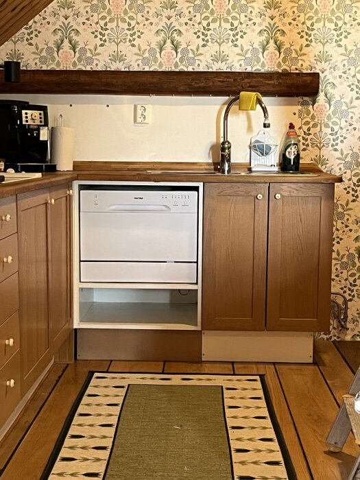 Diskmaskin installerad under köksbänk, omgiven av skåp, med en diskho och diskmedel ovanpå träbänken. Blommig tapet i bakgrunden.