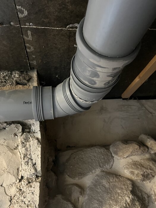 Grå avloppsrör med svängda kopplingar i en stenväggsmiljö.