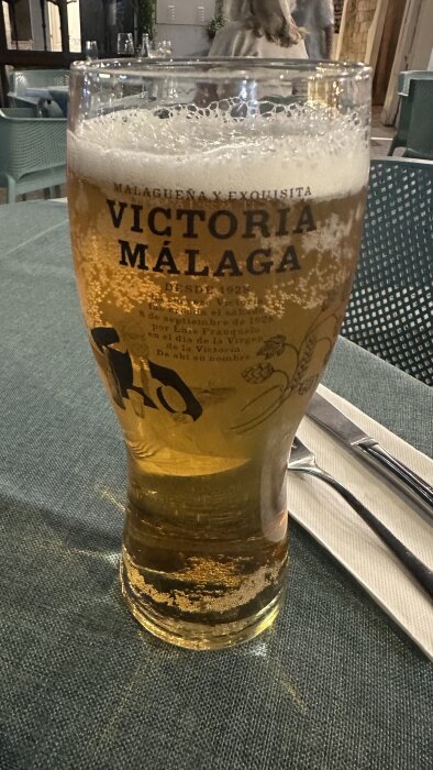Ölglas med texten "Victoria Málaga" på ett bord, perfekt för varma dagar.