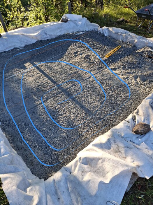 Paddad yta av grus med blå linjer som visar mönstret. Gränsen täckt av filt. Hjulbär med spade och vattenpass vid kanten.