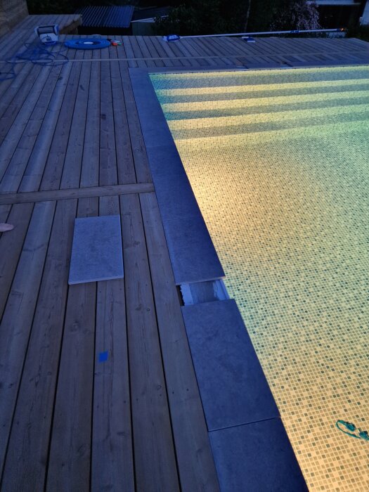 Trätrall vid pool med ojämn sarg. Stenarna vid sidan av poolen är inte jämna, vilket skapar luckor. Poolen är upplyst och bilden är tagen i skymningen.