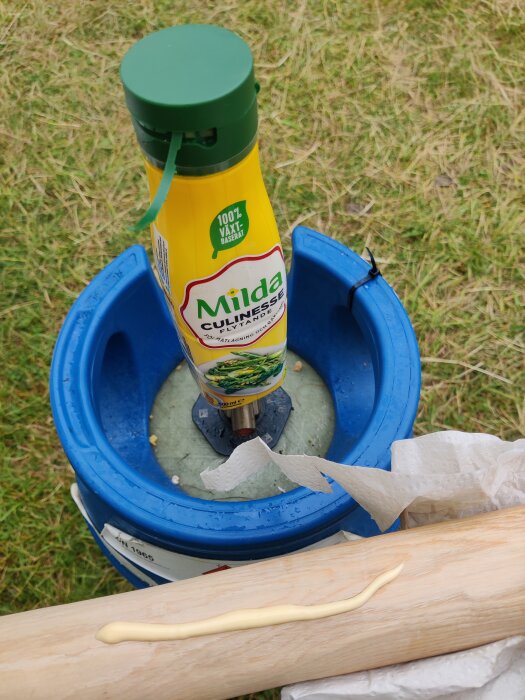 En flaska Milda Culinesse står i en blå behållare med gräs i bakgrunden. En träbit ligger med en gul kräm utspridd på ytan.