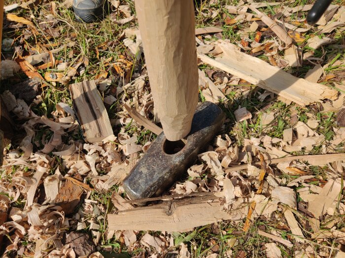 En yxa bredvid ett träskaft infogat i en slägga, omgiven av träspån på marken.