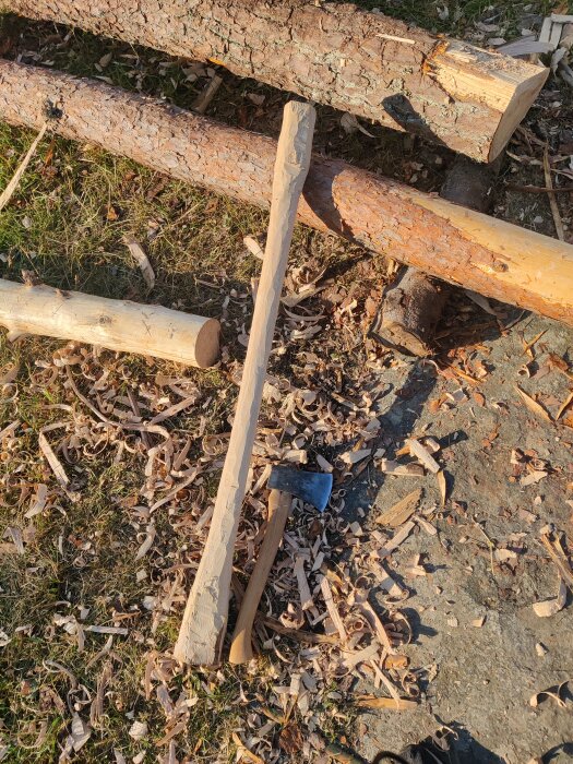Handgjort släggskaft av trä bredvid yxa och träflisor, omgiven av stockar utomhus.