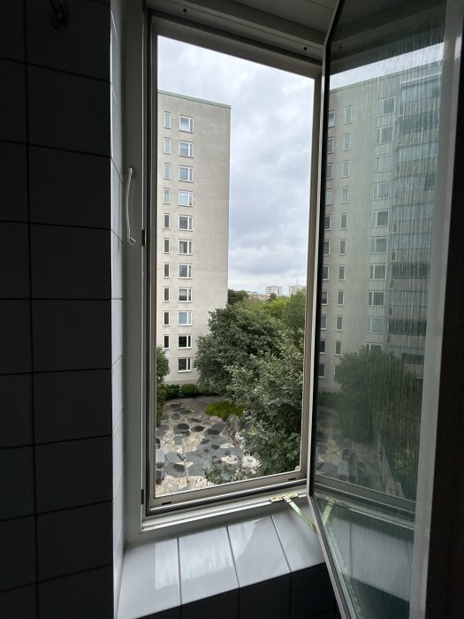 Öppet badrumsfönster sett inifrån med synliga kaklade väggar och en utsikt mot ett grönområde och flerbostadshus utanför.