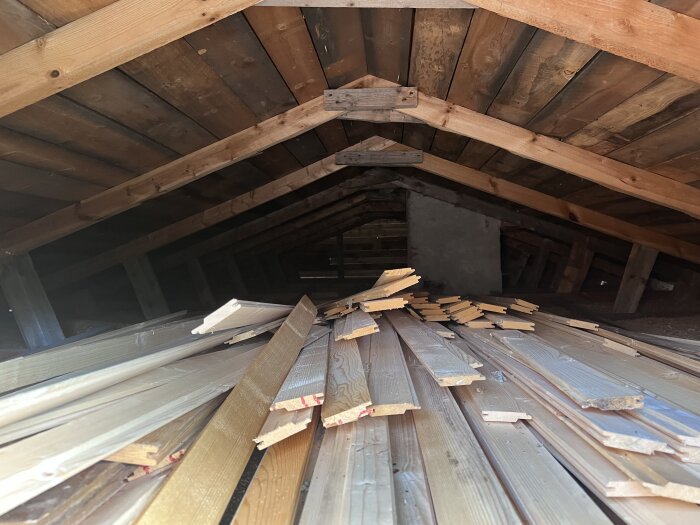 Ett loftutrymme med lågt tak och synliga träbalkar samt liggande brädor staplade på varandra, som en del av ett renoveringsprojekt.