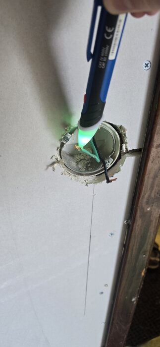 Elektrikertestare med grönt ljus mot modifierad väggdosa; exponerade svarta och gröna trådar syns, osäkerhet kring installationens funktion.