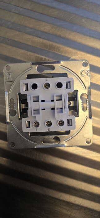 En strömbrytare med metallram och plastfront, märkt med L och N, ligger på ett ljusbrunt ytmönster.