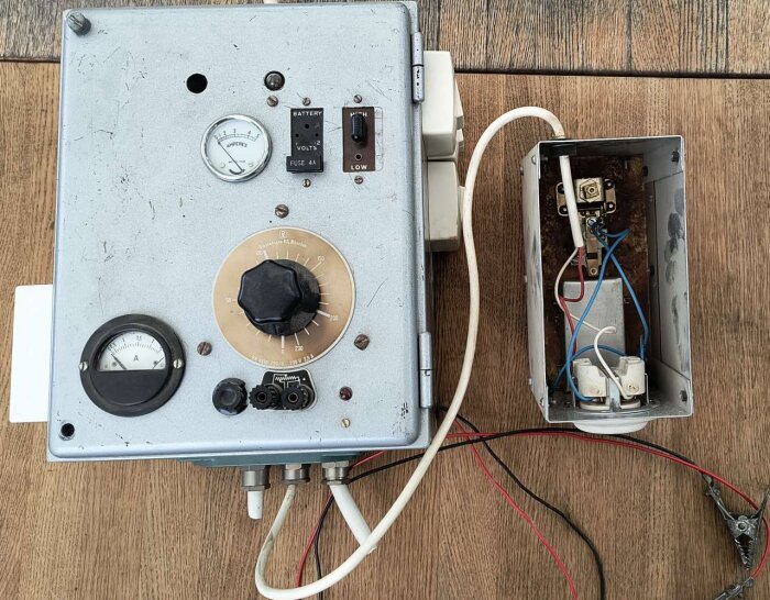 En gammal elektrisk apparat med mätarmetrar, ratt och uttag, troligen från 1950-talet, placerad på ett trägolv.