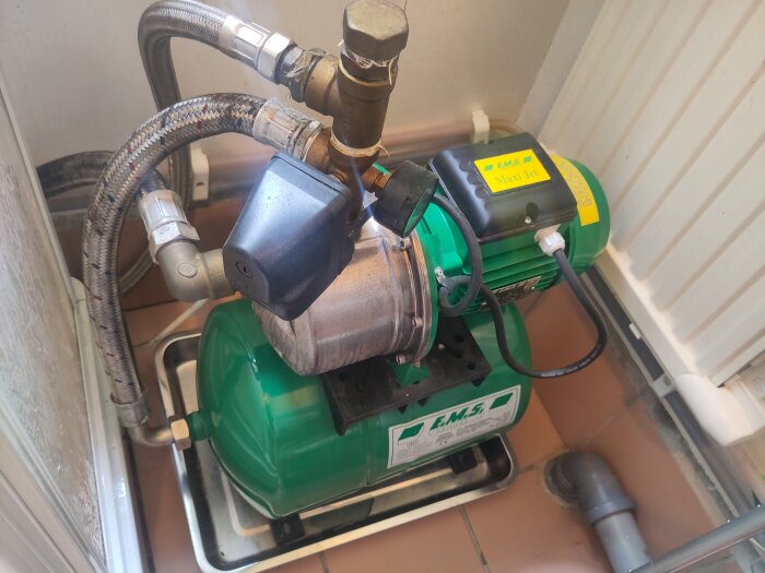 Grön jetpump med flera kopplade slangar, hydrofor och motor placerad i ett hörn av ett rum med kakelgolv.