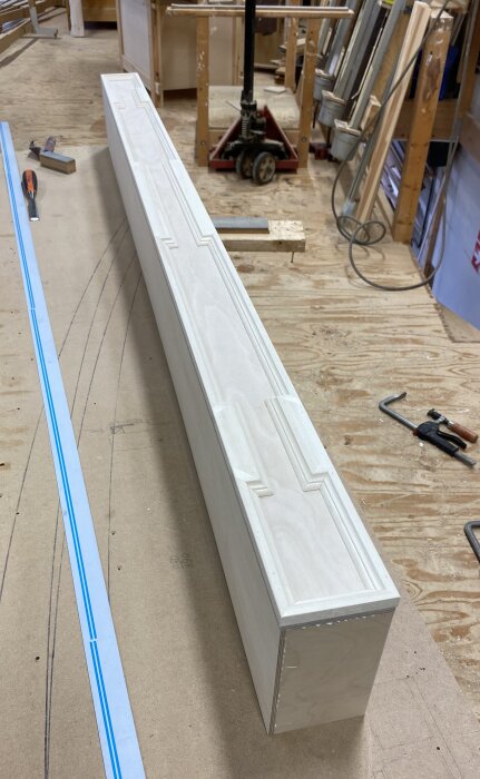 Träpanel med dekorativ profil på golvet i en verkstad, omgiven av verktyg och andra material.