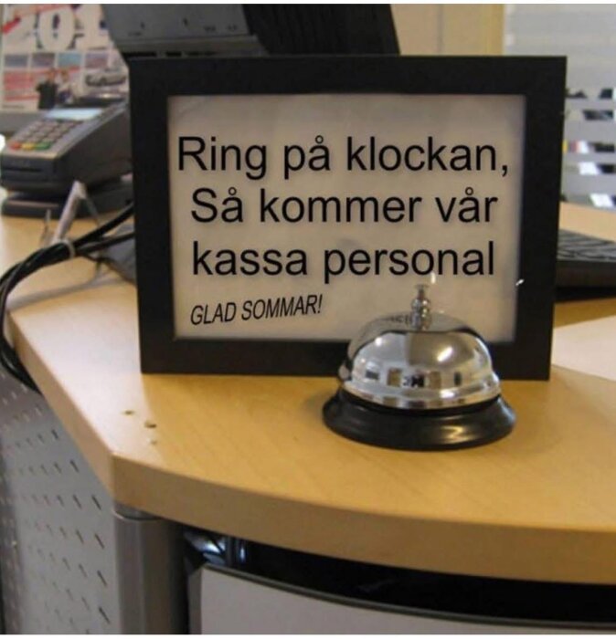 Skylt på disk med text "Ring på klockan, Så kommer vår kassa personal, Glad sommar!" och en ringklocka framför.