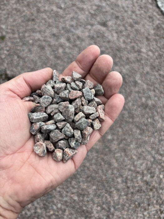 En hand som håller en näve gårdgrus av olika storlekar mot en bakgrund av mer grus.