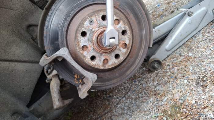 Bromsskiva med rost, en skiftnyckel hålls mot navet, omgiven av bromsok. Bilen är upppallad med en domkraft på grusunderlag.