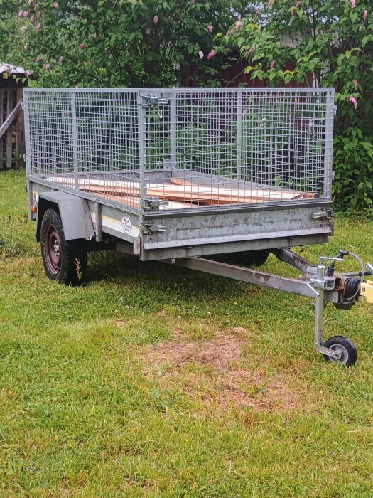 En släpvagn med ett måttillverkat galler parkerad på en gräsmatta, omgiven av gröna buskar och blommor i bakgrunden.