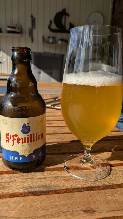 Flaska och glas med St. Feuillien Tripel-öl på ett träbord, solbelyst bakgrund.