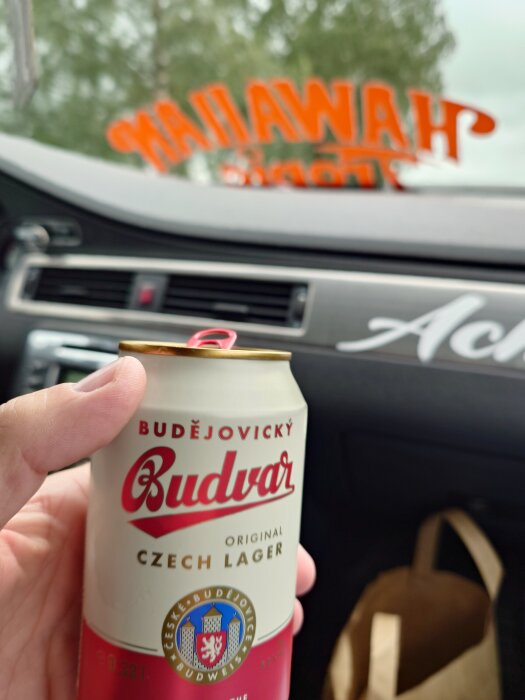 Hand håller en burk Budějovický Budvar tjeckisk lager i en bil, med text på rutan i bakgrunden.