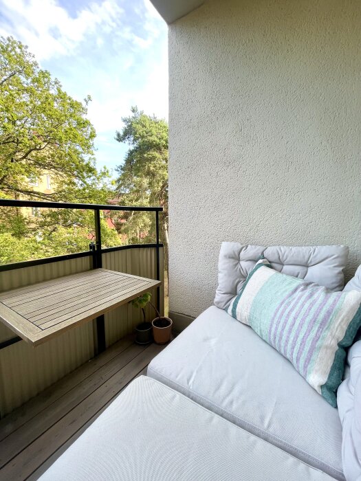 Balkong med vit putsad vägg, trädgårdsmöbler, och två krukväxter på trägolv. Fantastisk utsikt över grönskande område.