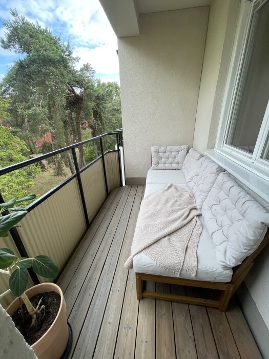 Balkong med träplankgolv, beige soffgrupp med kuddar, filt och en krukväxt intill ett räcke, med träd i bakgrunden.