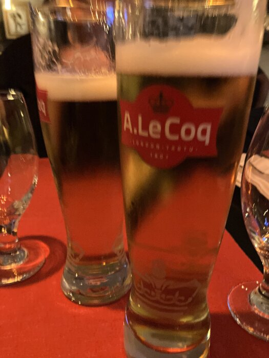 Två glas med öl på ett bord med röd duk, etikett med text "A. Le Coq" synlig.