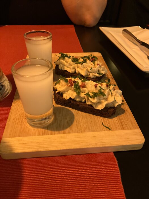 Bröd med toppings och persilja på en träbricka bredvid två glas med vit dryck.